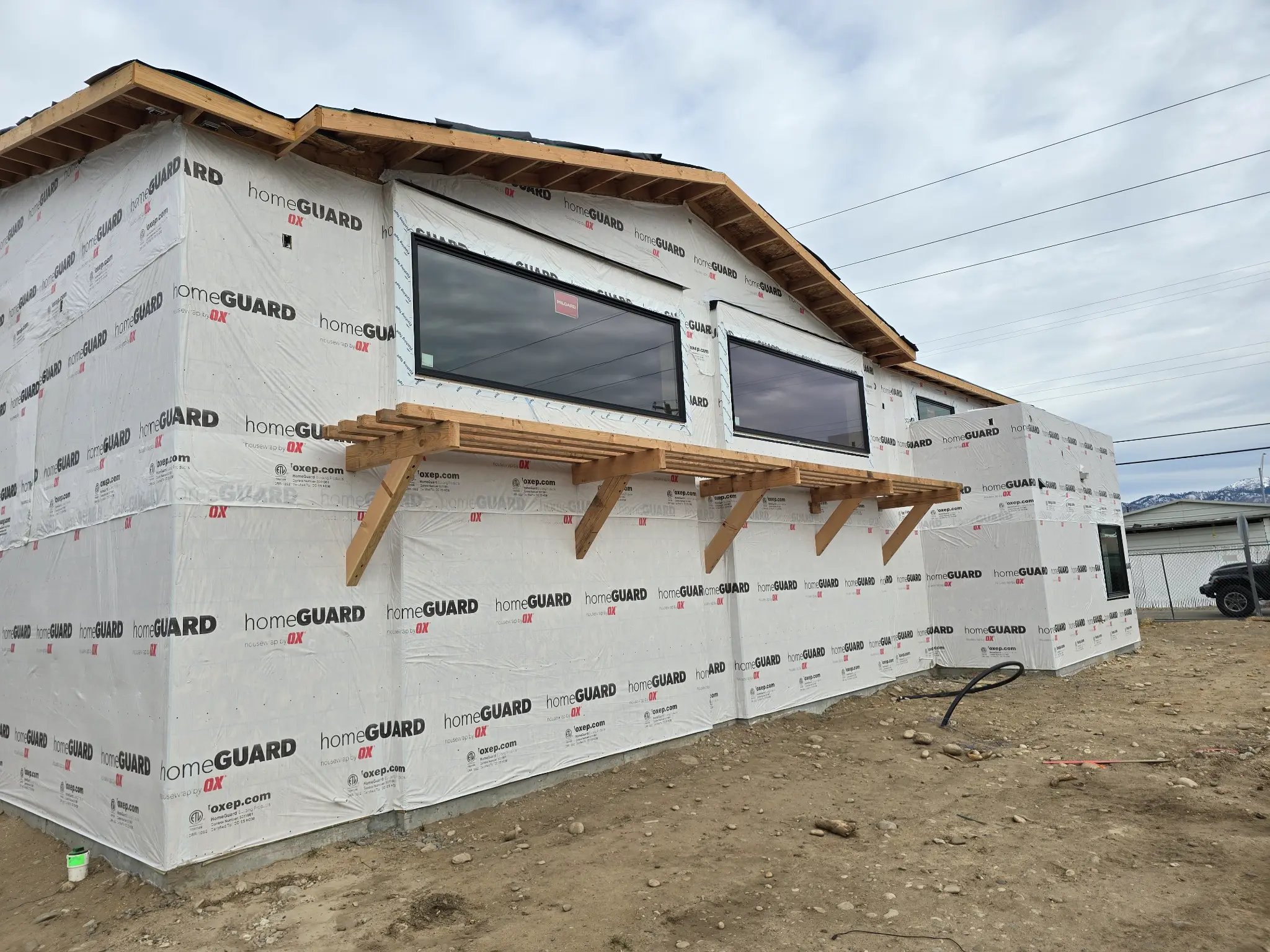 Before photo of commercial building office wing side wall wrapped in HomeGuard housewrap, showing 3 panoramic windows and wood canopy bracket framing