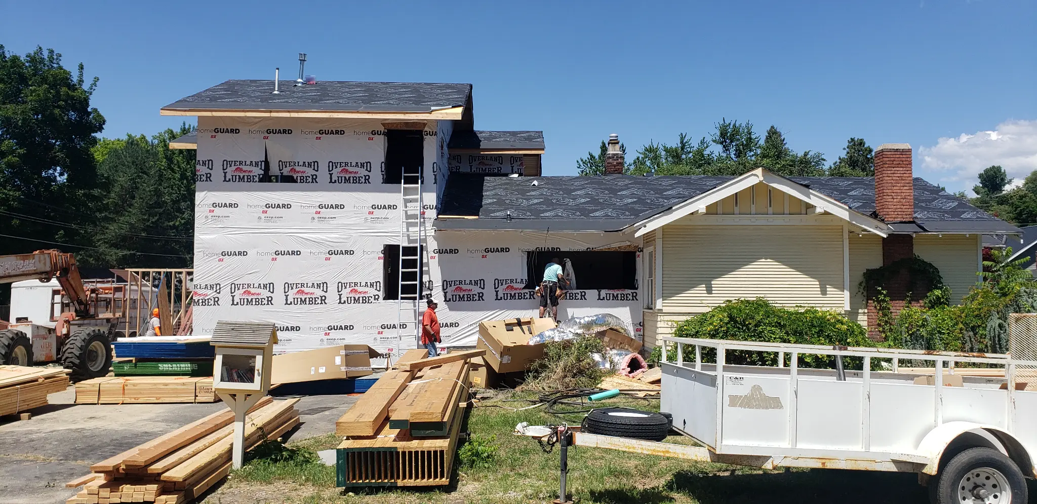Before photo of House 2 during HBSI exterior work with housewrap, roofing prep, ladders, and construction materials on site